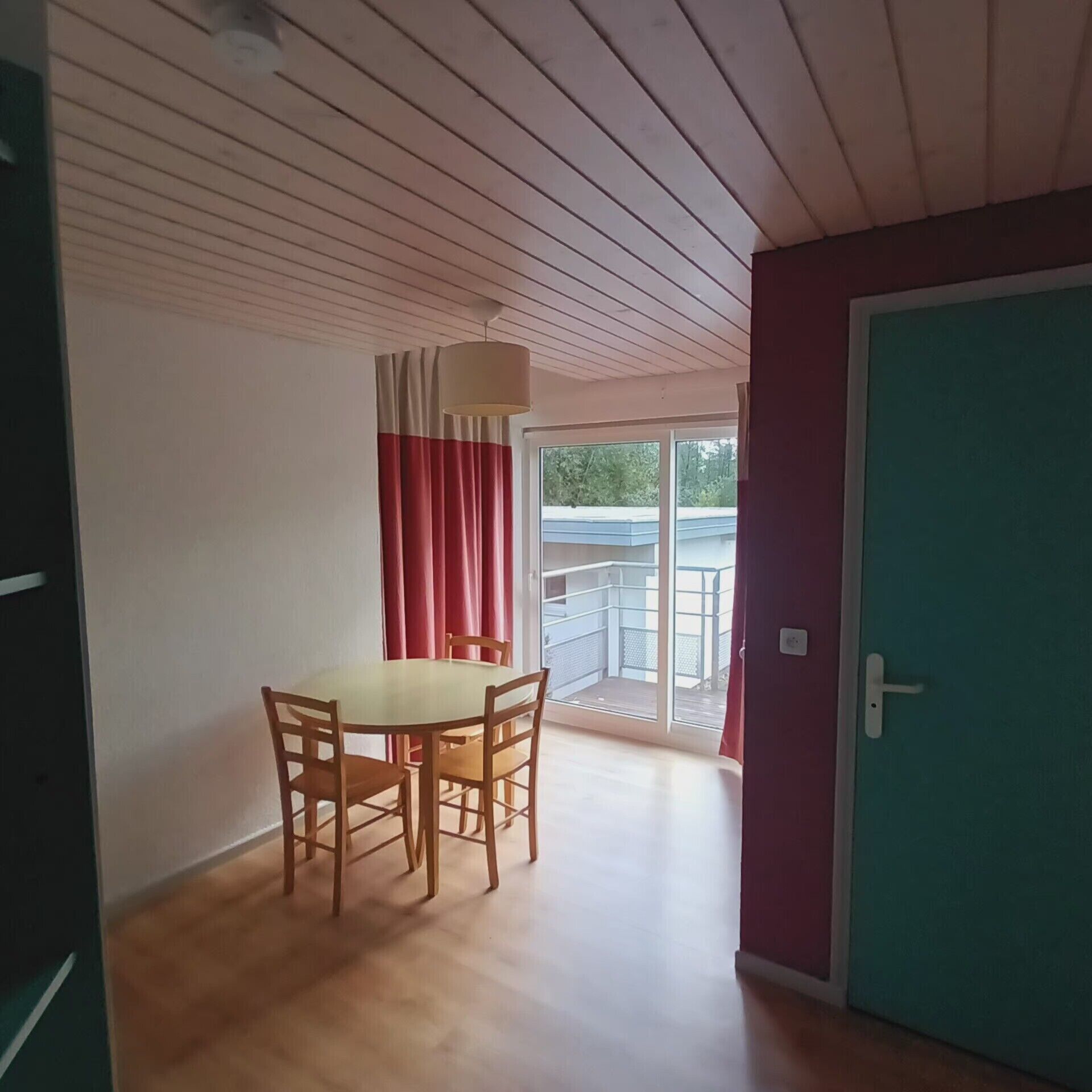 Une pièce comprenant une table à manger et des chaises, un ventilateur de plafond, une porte-fenêtre coulissante donnant sur un balcon et une porte verte.