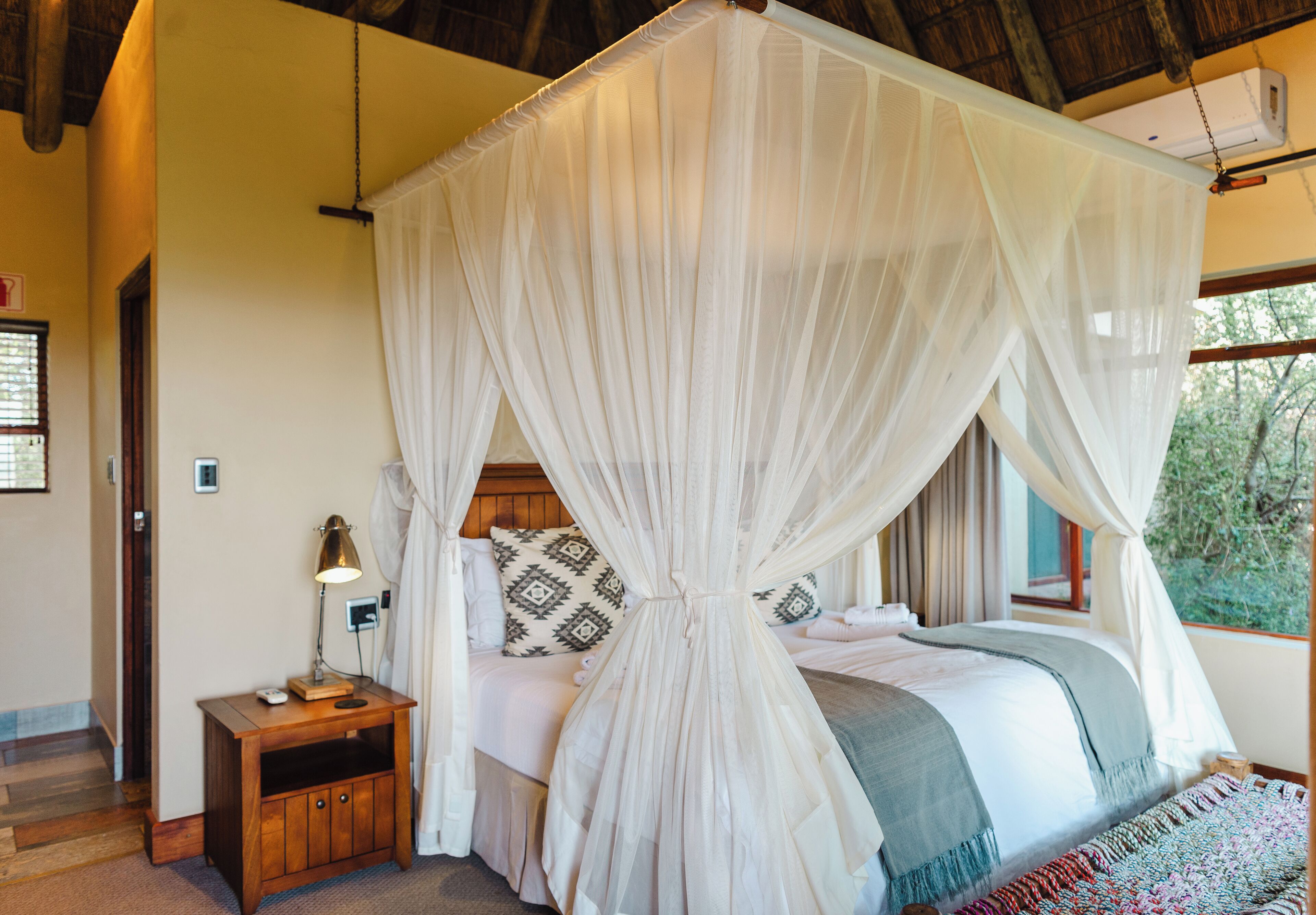 A bedroom with a four-poster canopy bed, a bedside table with a lamp, and a view of greenery outside.