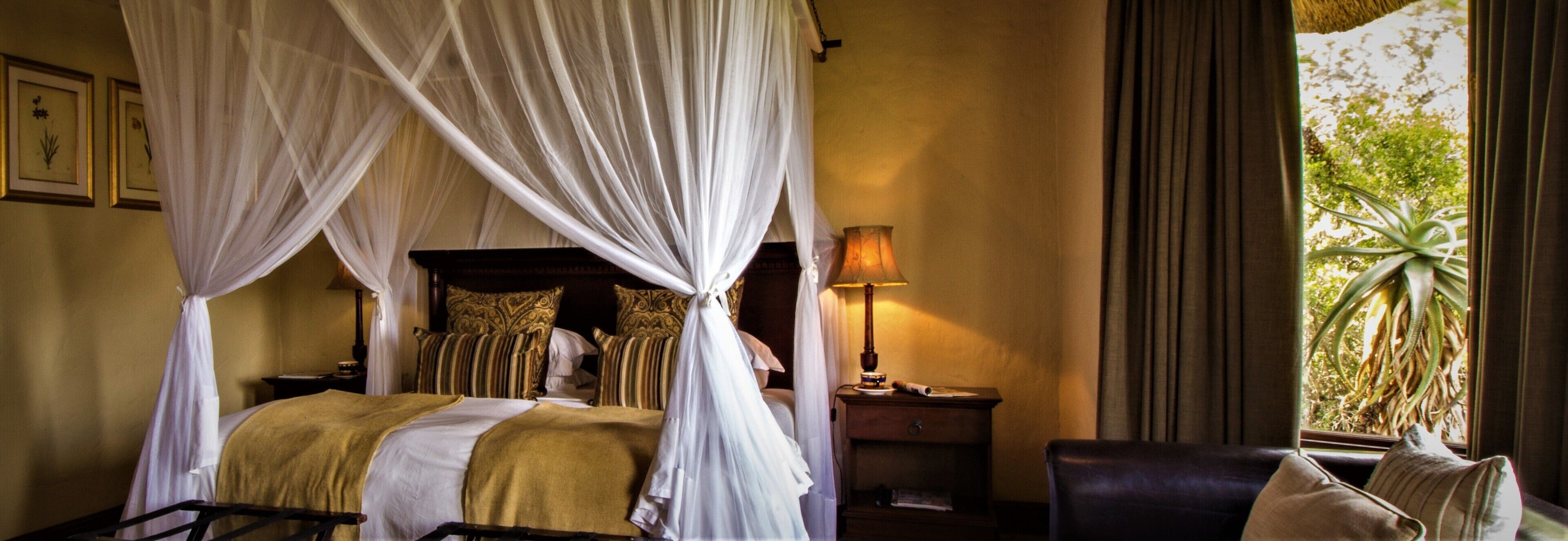 A four-poster bed with a canopy, a bedside table with a lamp, a chair, and a view of greenery outside.