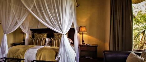 A four-poster bed with a canopy, a bedside table with a lamp, a chair, and a view of greenery outside.