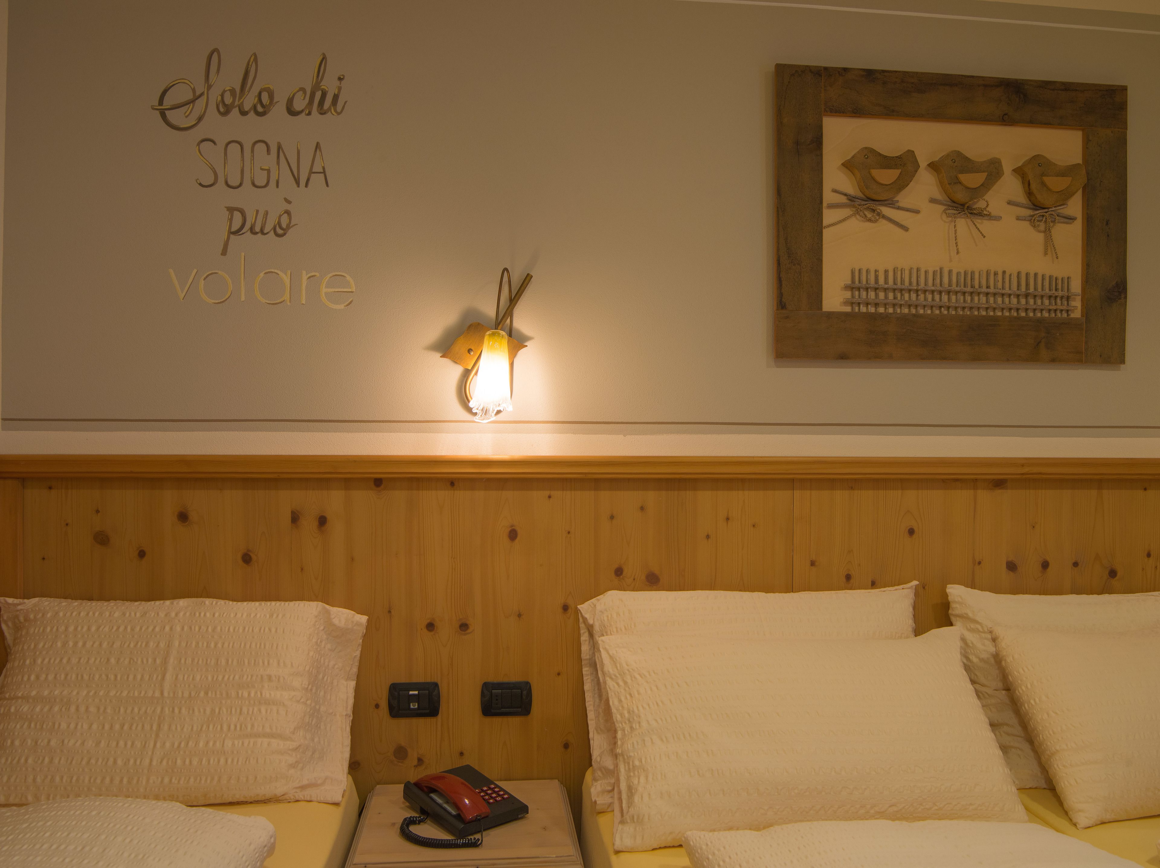Une chambre d’hôtel avec une tête de lit en bois, une lampe fixée au mur, une œuvre d’art encadrée représentant des oiseaux et un panneau avec du texte en italien.