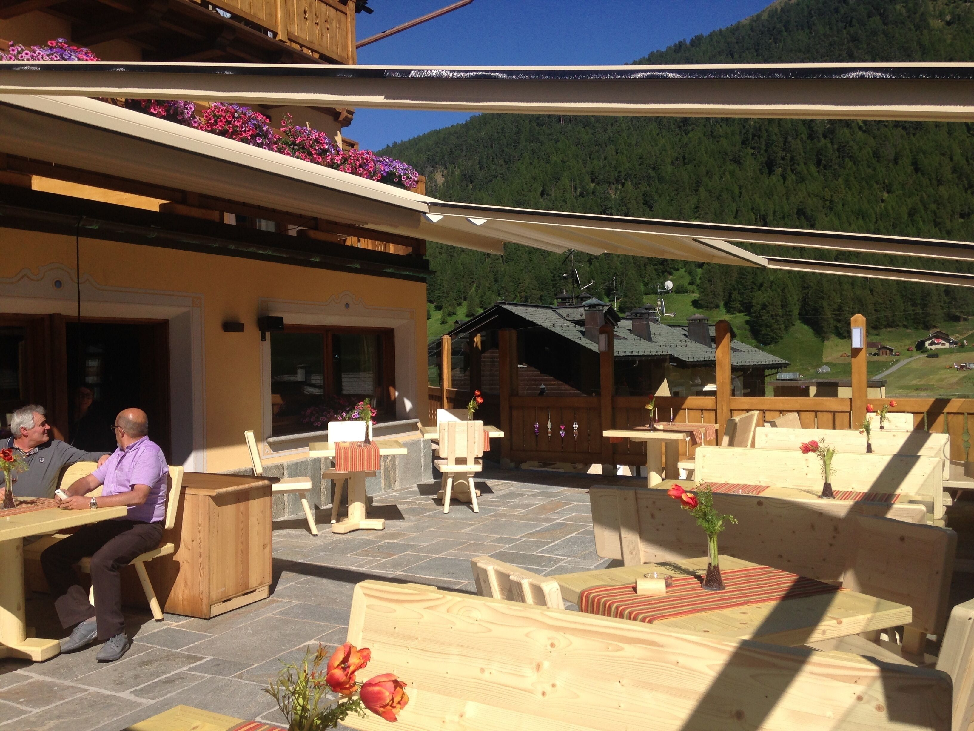 An outdoor seating area with wooden tables and chairs, a stone floor, and flower arrangements.