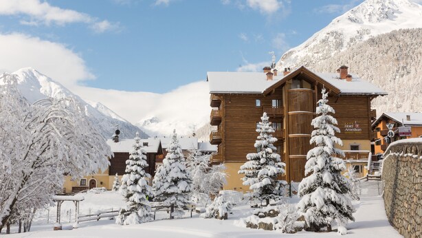 A wooden lodge with a snow-covered roof, surrounded by snow-laden trees and mountains in the background. - Hotel Amerikan (Livigno)