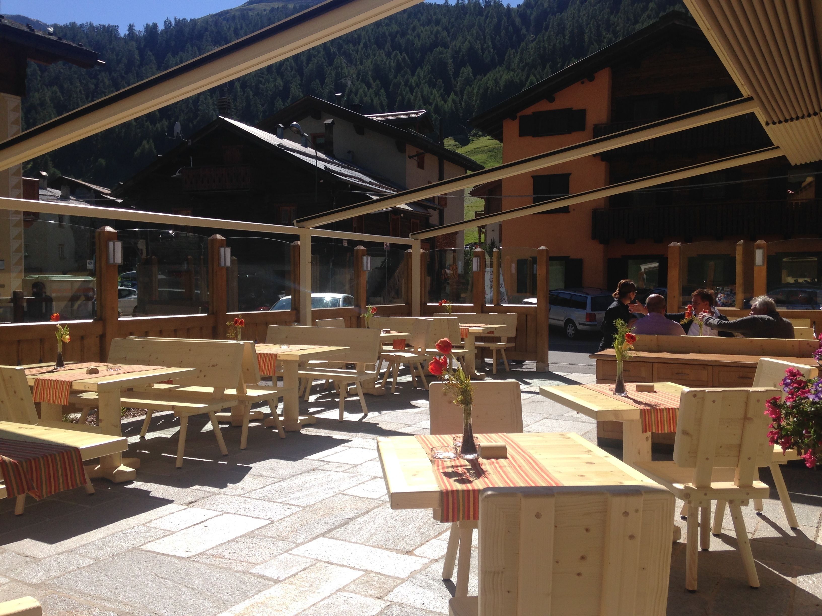 An outdoor seating area with wooden tables and chairs, a stone floor, and a mountain backdrop.