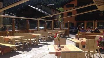 An outdoor seating area with wooden tables and chairs, a stone floor, and a mountain backdrop.