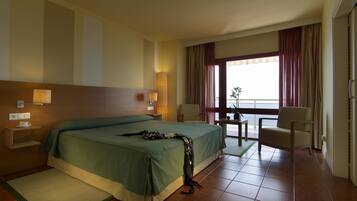 Ein Hotelzimmer mit einem großen Bett, einem Nachttisch mit Lampe, einem Sessel, einem kleinen Tisch und Blick ins Freie.