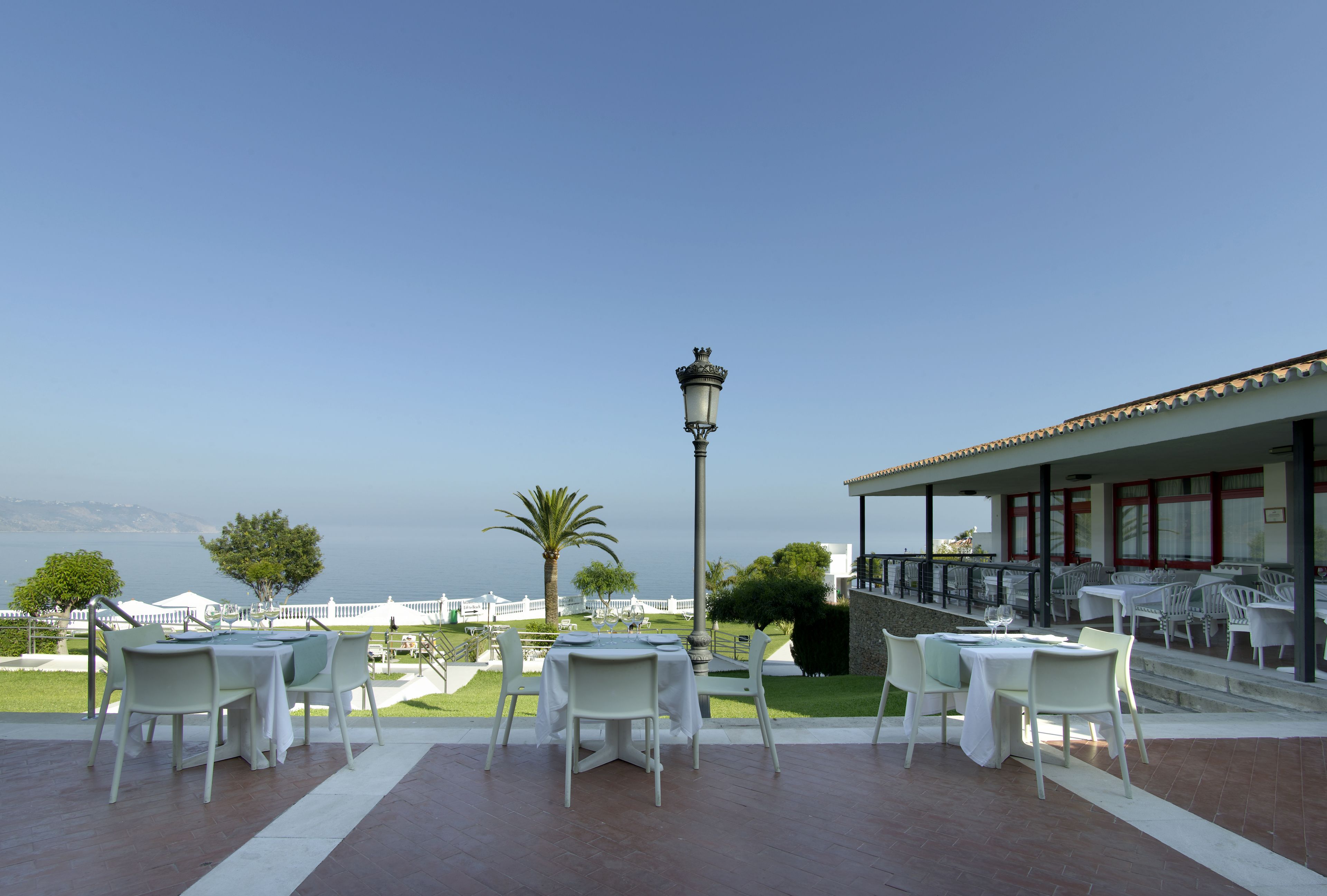 Un área de comedor al aire libre con mesas y sillas blancas, un farol y vista al mar.