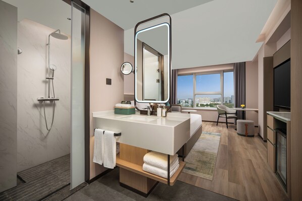 Un moderno baño de hotel con ducha, un espejo grande, tocador con lavamanos y vista a la ciudad a través de un ventanal.