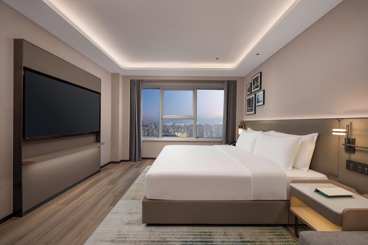 Une chambre d’hôtel moderne dotée d’un grand lit, d’une télévision à écran plat, d’une vue sur la ville et d’une table de chevet avec une lampe.