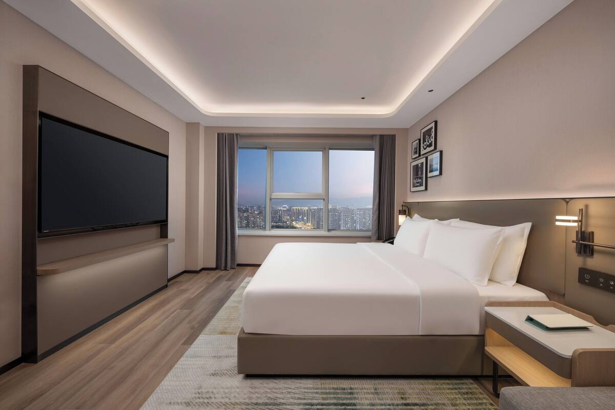Habitación de hotel moderna con una cama grande, un televisor de pantalla plana, vista a la ciudad y una mesita de noche con lámpara.