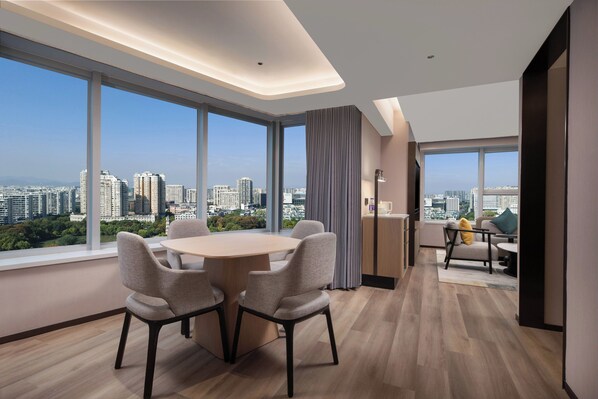 A modern hotel room with a large dining table, comfortable seating, and a city view through expansive windows.