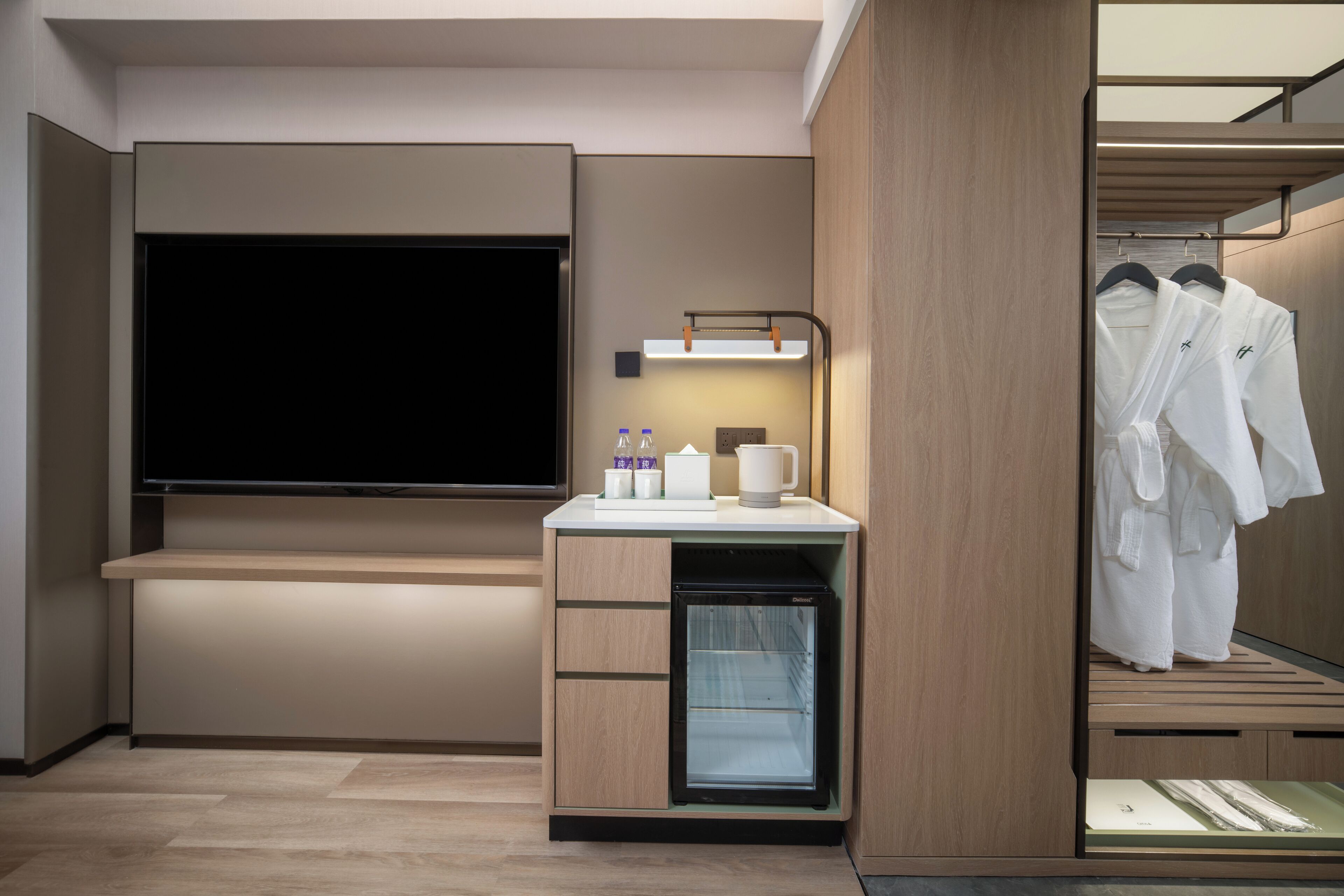 A modern hotel room with a flat-screen TV, a mini-fridge, a vanity with a mirror, and a walk-in closet with hanging robes.