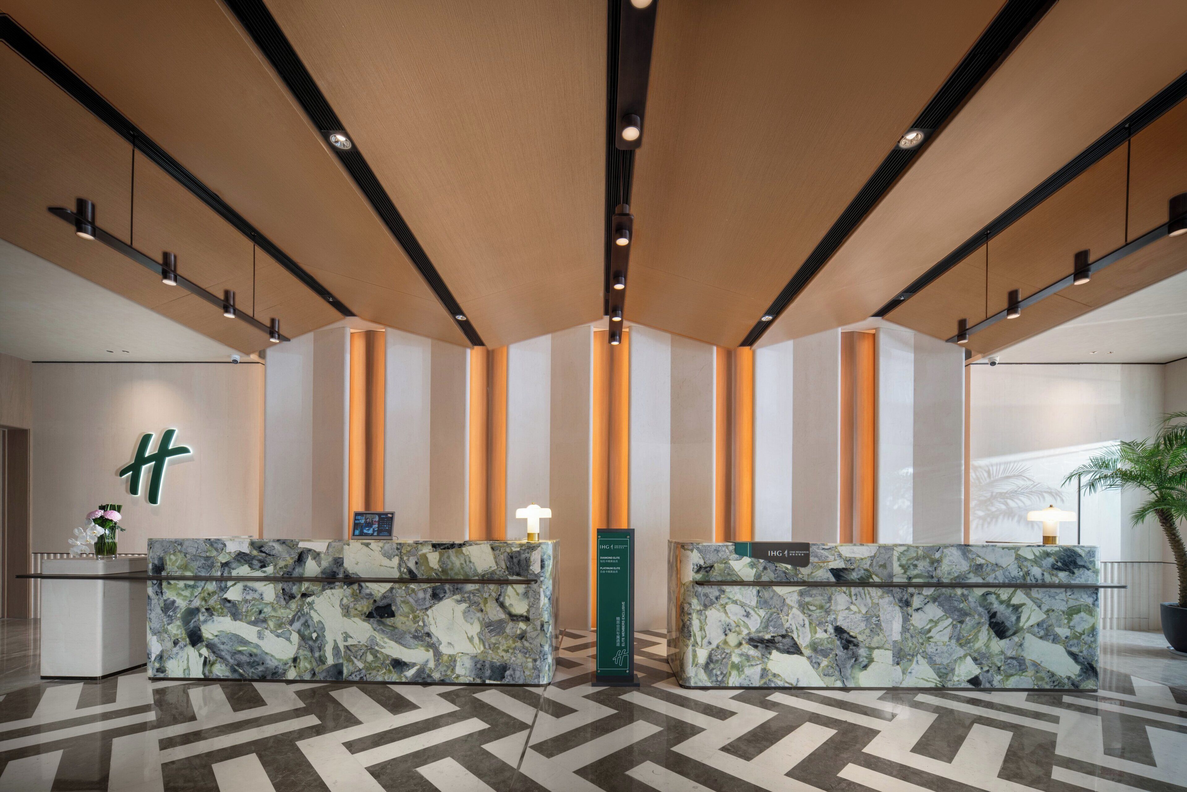 Ein Hotel-Rezeptionsbereich mit Marmor-Rezeptionstheken, einem großen grünen Schild und einem geometrisch gemusterten Boden.
