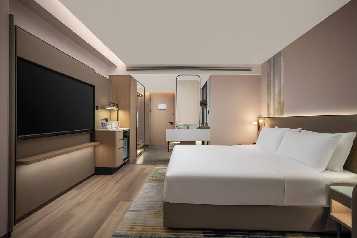 Una habitación de hotel moderna con una cama grande, un televisor de pantalla plana, un escritorio y un baño con espejo y lavabo.