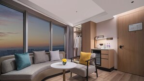 Ett modernt hotellrum med en böjd soffa, ett litet runt bord med en vas med blommor, ett litet kök och ett stort fönster med utsikt över staden.