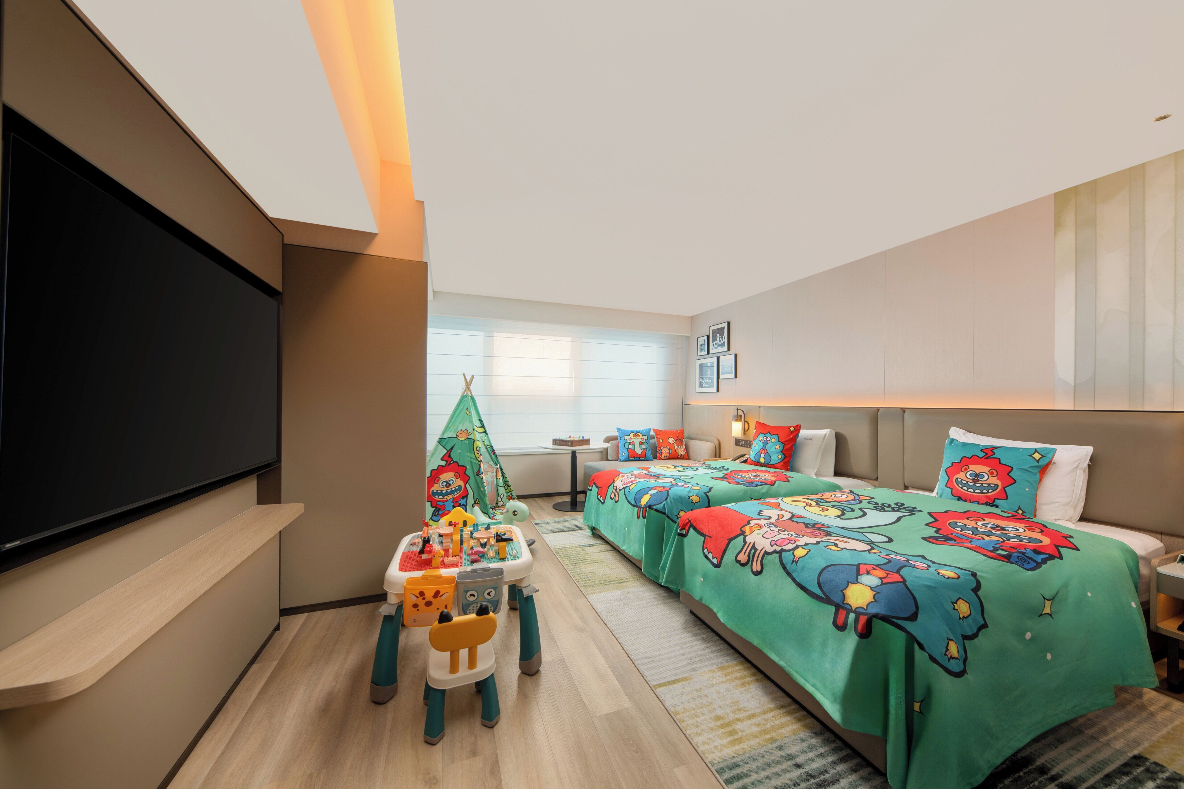 Een hotelkamer met een grote flatscreen-tv, een bed met kleurrijk beddengoed, een speelhoekje met speelgoed en een raam met zonwering.