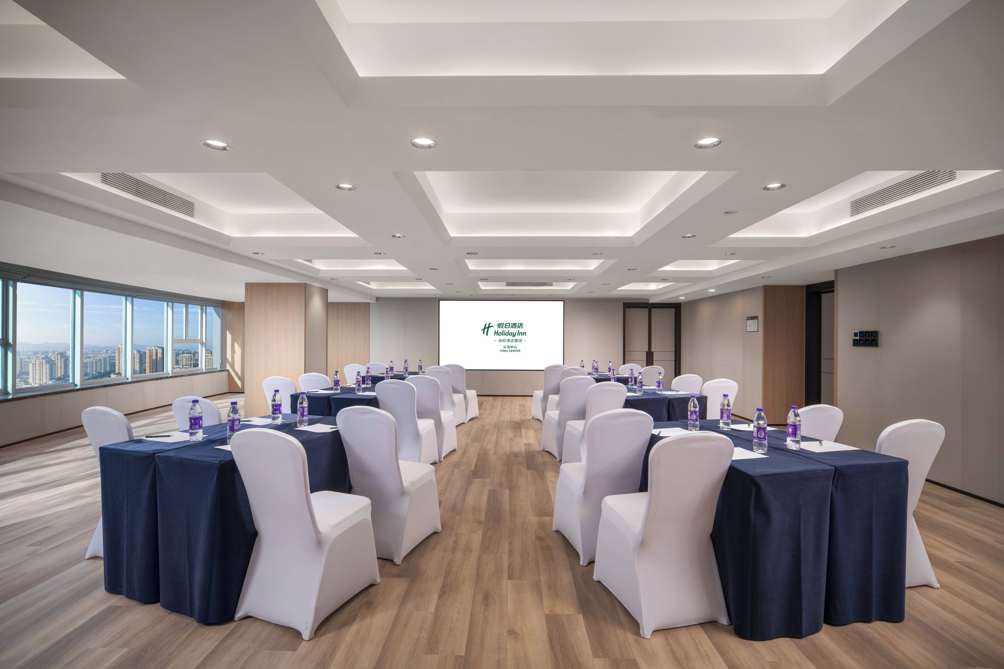 Un salón de conferencias con mesas dispuestas para una reunión, botellas de agua y una pantalla para presentaciones al frente.