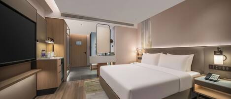 A modern hotel room with a large bed, bedside tables, a flat-screen TV, a mirror, and a bathroom visible through an open door.