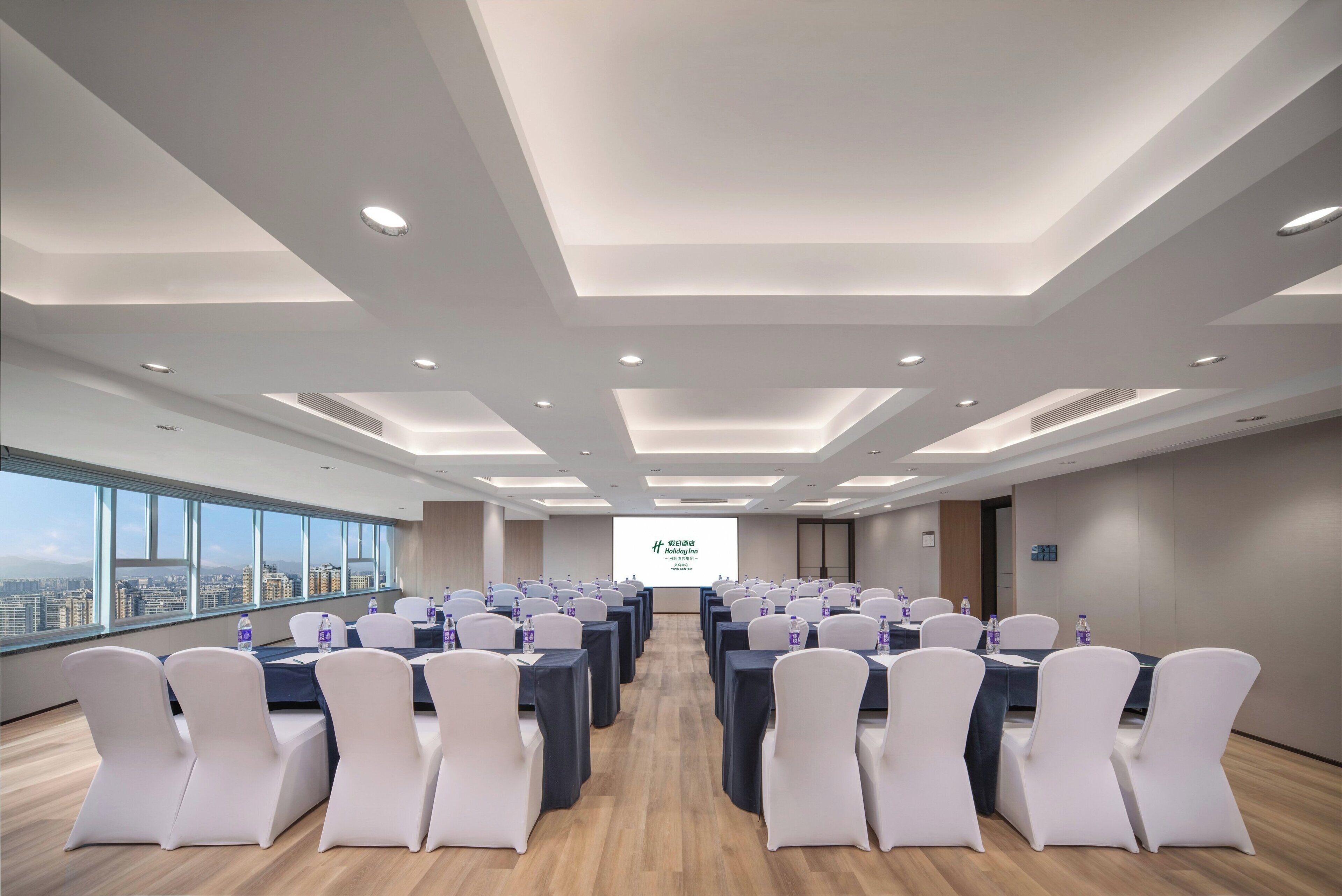 A conference room with tables and chairs, a projector screen, and a city view through large windows.