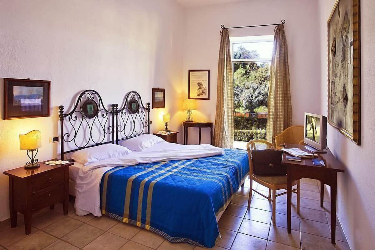 Ein Schlafzimmer mit Bett, Nachttischen, Schreibtisch und Stuhl. Vorhänge, ein Fenster und ein Bild an der Wand sind ebenfalls vorhanden.