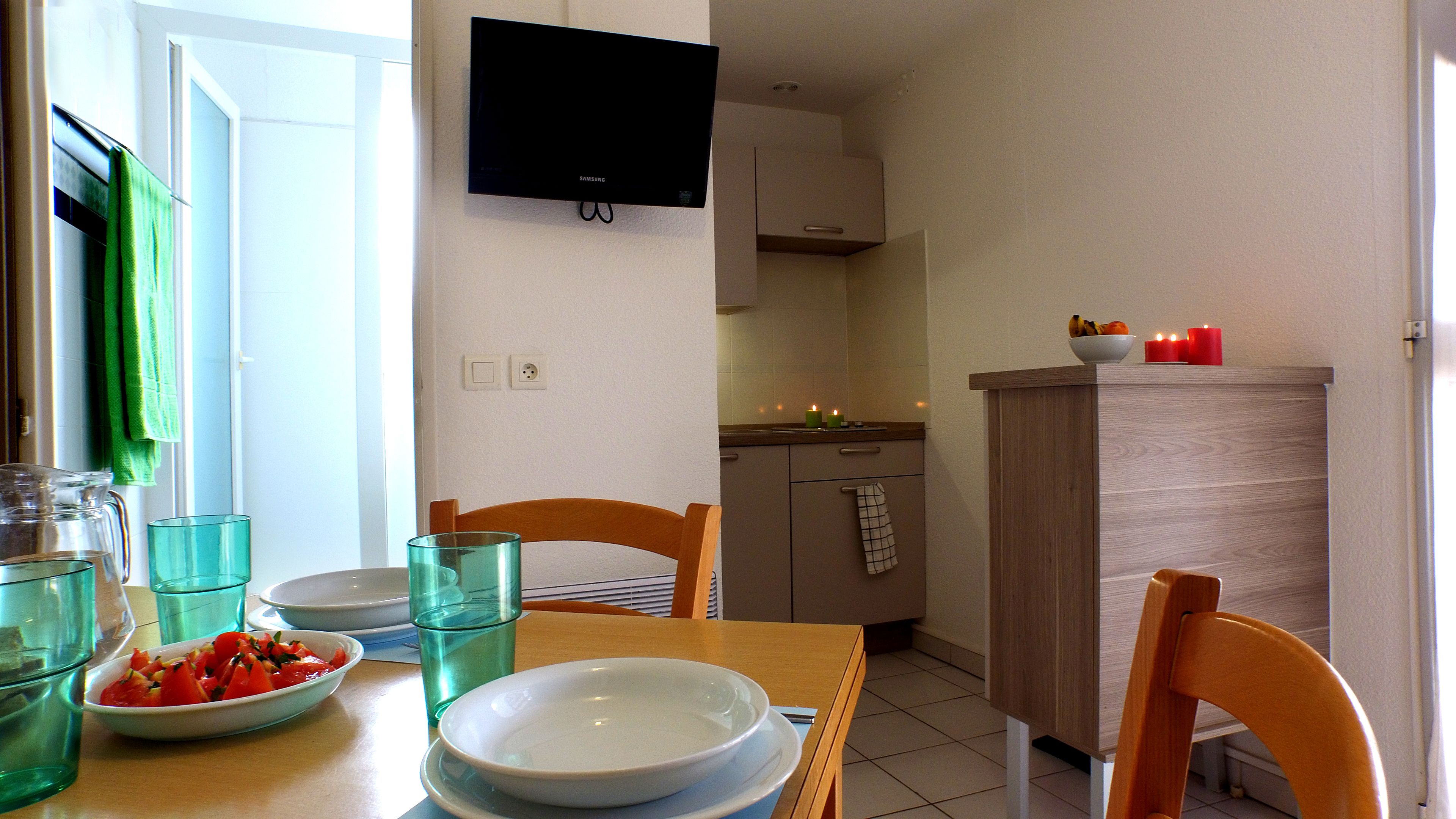Un coin repas avec une table dressée pour deux personnes, un téléviseur fixé au mur et une cuisine en arrière-plan.