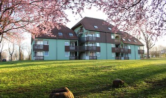 Un edificio residencial de varios pisos con balcones, rodeado por una zona ajardinada y árboles de cerezo.