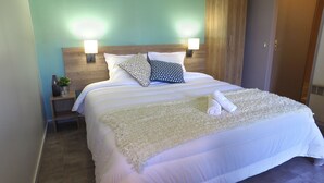 A neatly made bed with white linens, two patterned pillows, a white throw blanket, and a rolled towel on the bed. There are two bedside lamps on wooden nightstands with a small vase and a plant. - VVF Plaine d'Alsace Obernai Strasbourg (Obernai)