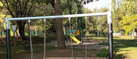 A playground with swings and a slide, surrounded by trees and gravel.