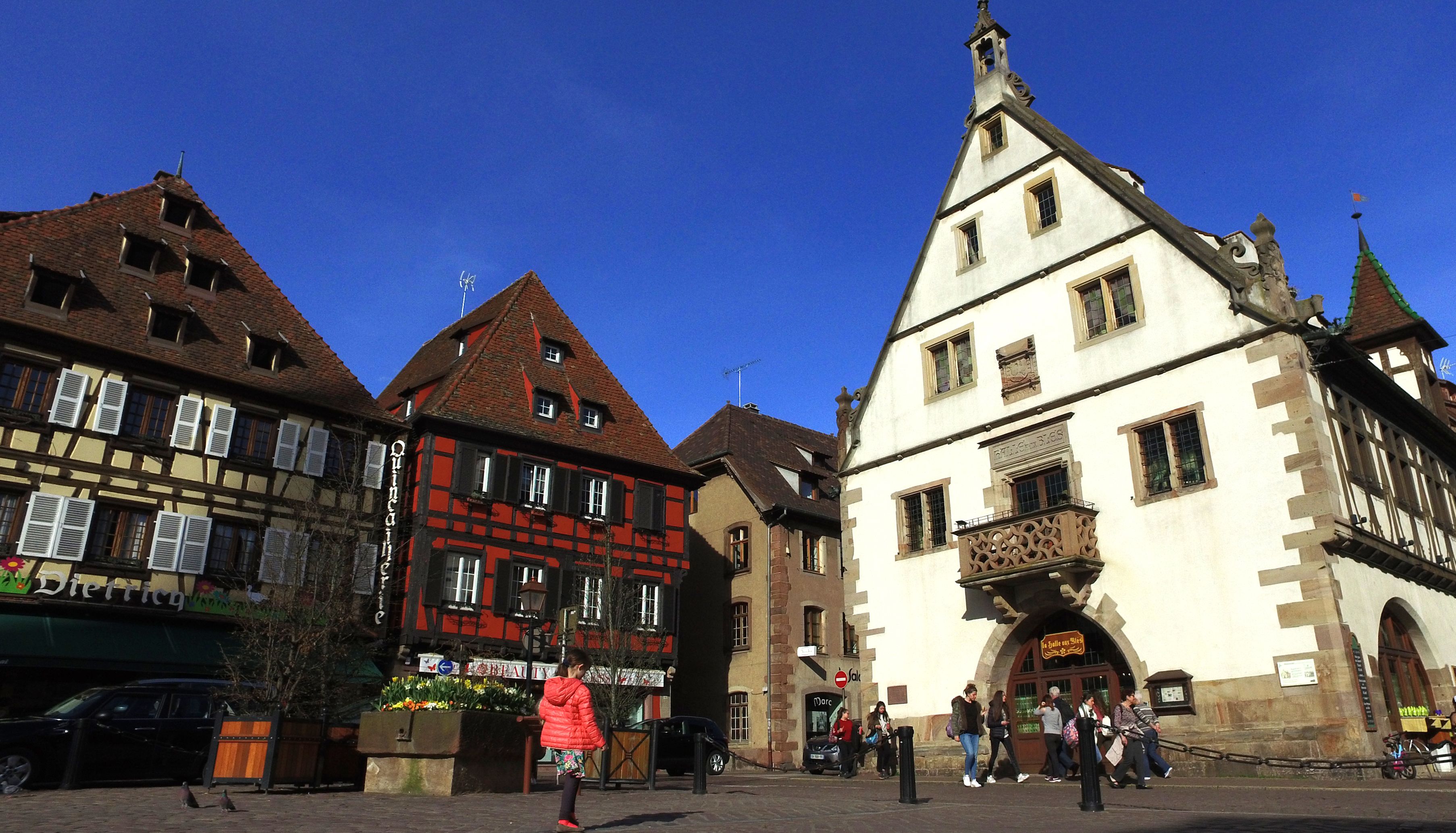 Een historisch Europees stadsplein met traditionele vakwerkhuizen, een opvallende kerk met een torenspits, en voetgangers.