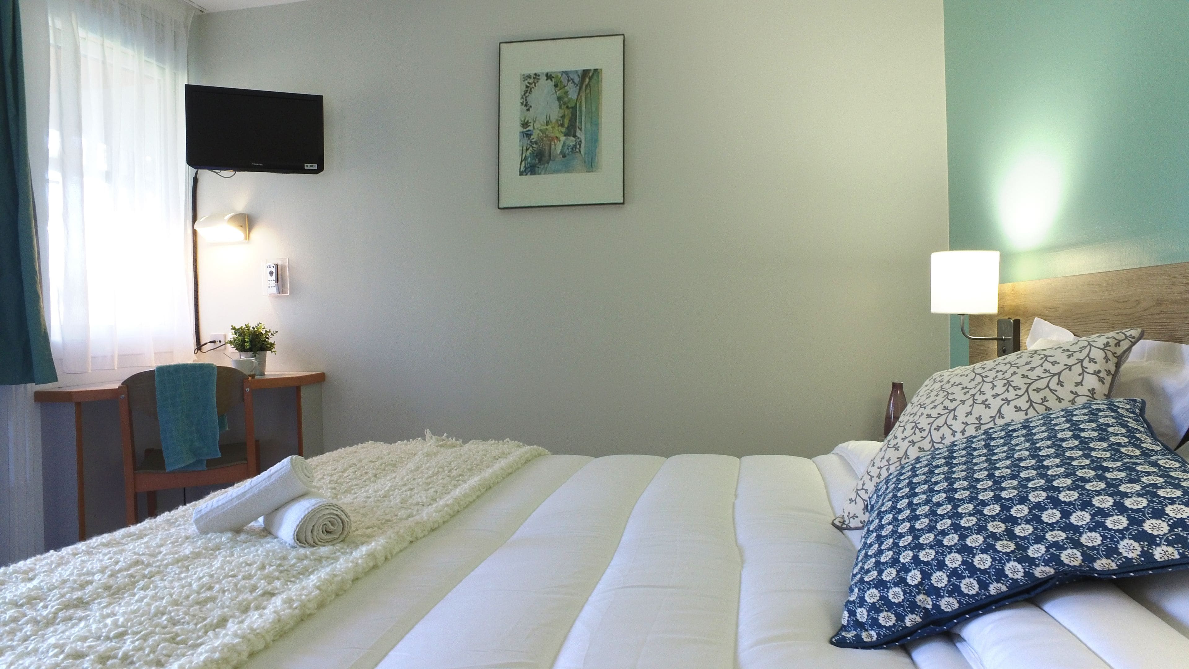 A hotel room with a bed, a desk, a chair, a TV, a painting, and a lamp.