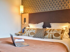 Quarto de hotel com cama, um laptop sobre a cama, mesa de cabeceira com abajur e parede estampada.