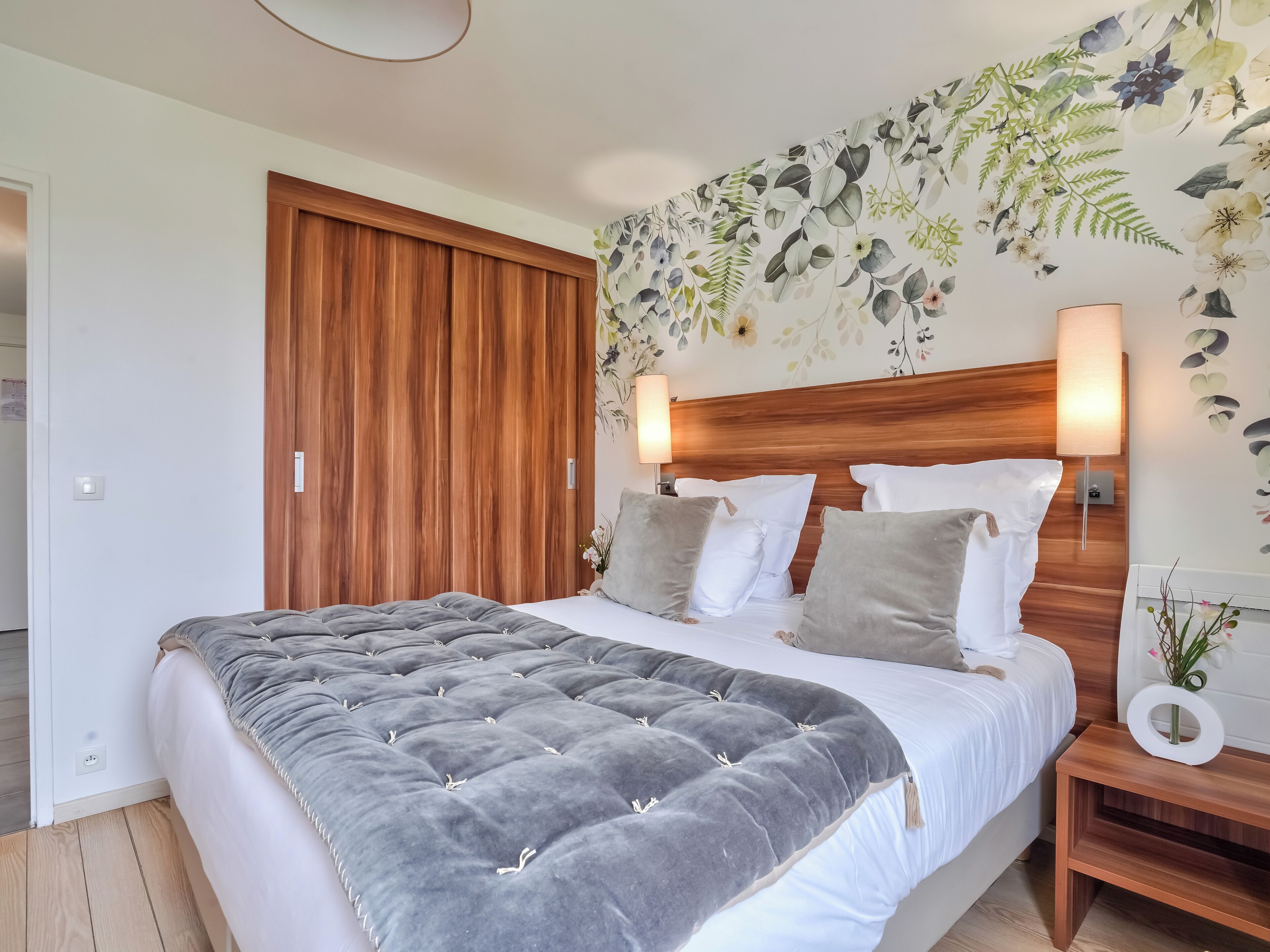 A bedroom with a large bed, wooden headboard, two bedside lamps, and a floral wall mural.