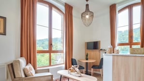 A cozy room with a rattan sofa, a round table, and a view of greenery through large windows.