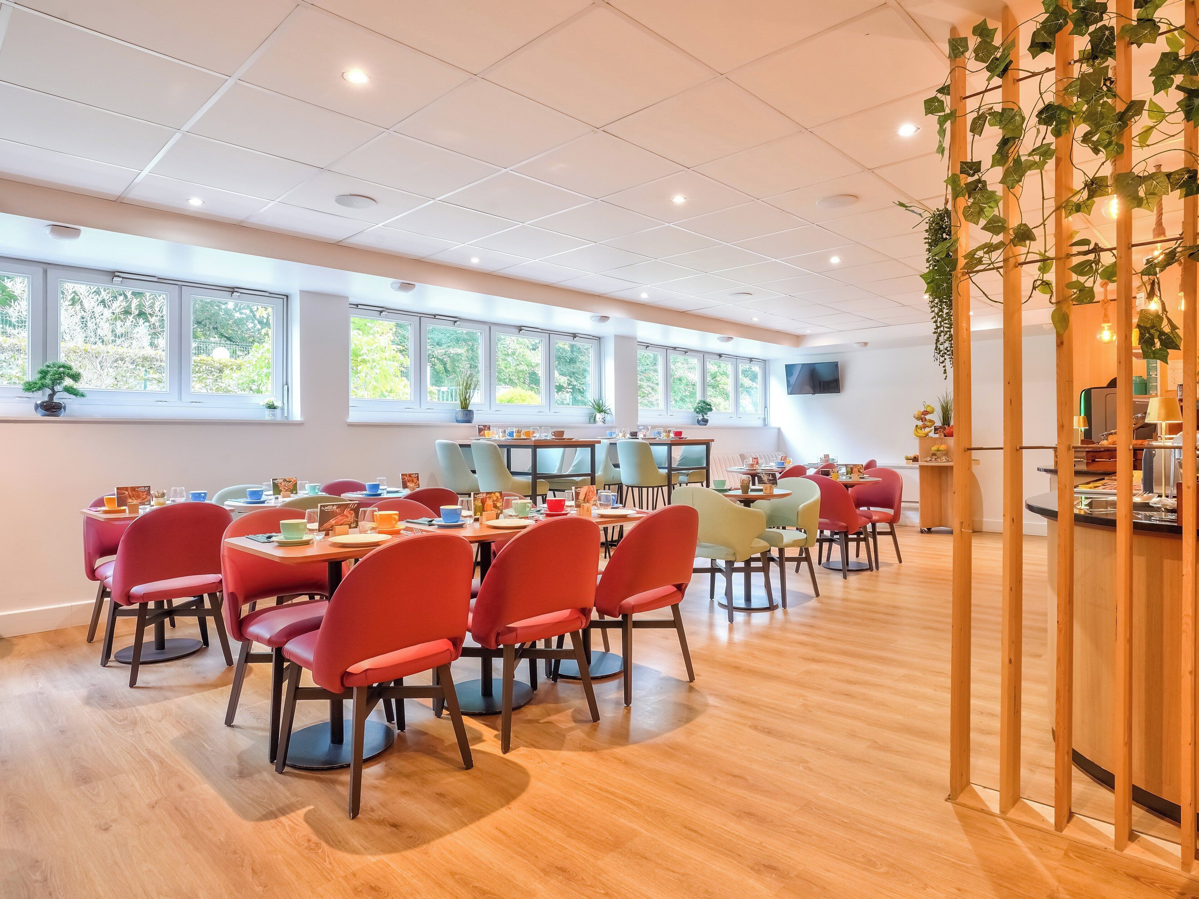 A spacious dining area with multiple tables and chairs, large windows with natural light, and a wooden partition with greenery.