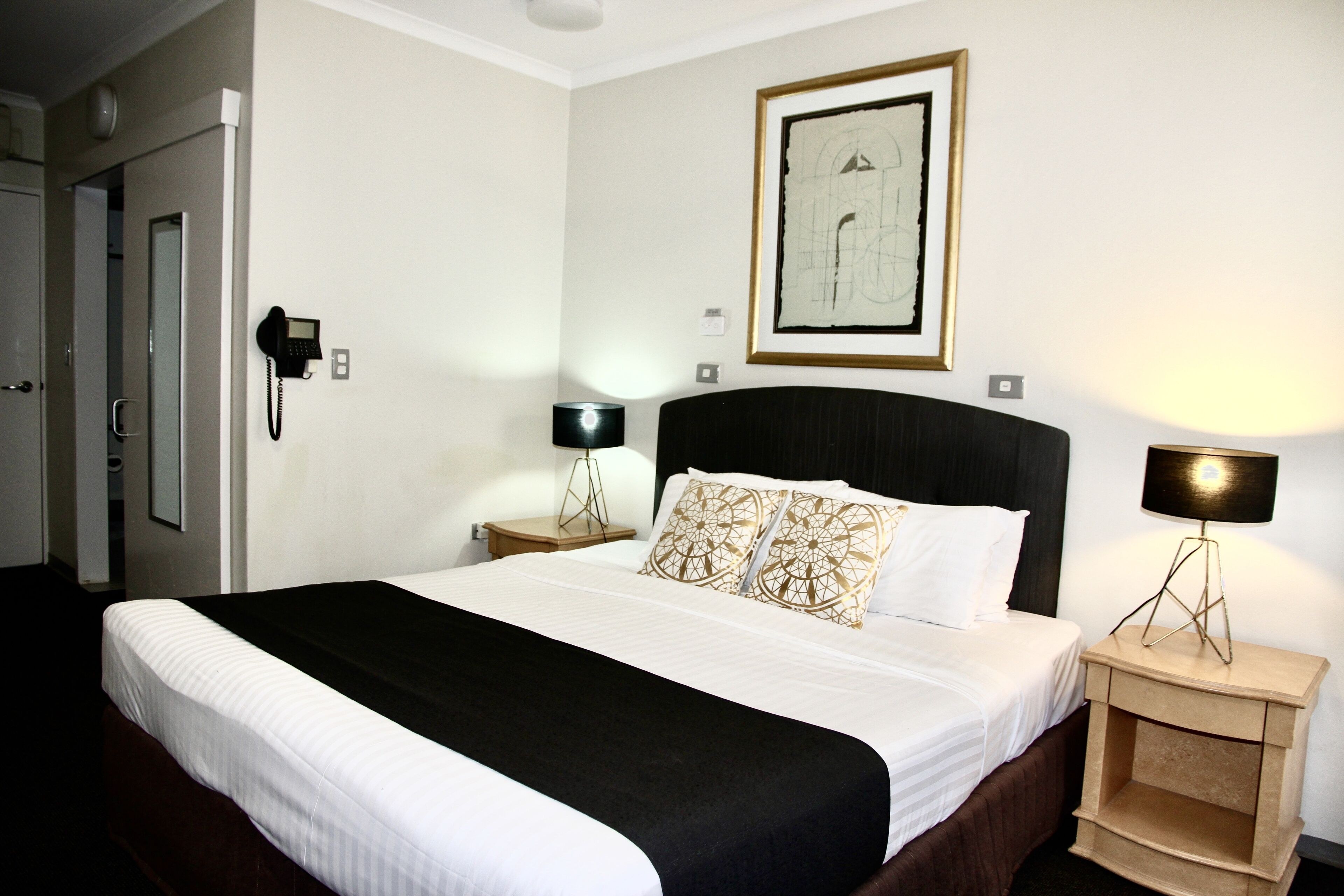 A neatly made bed with a black and white bedspread, a headboard with decorative pillows, a bedside lamp, and a framed abstract painting on the wall.