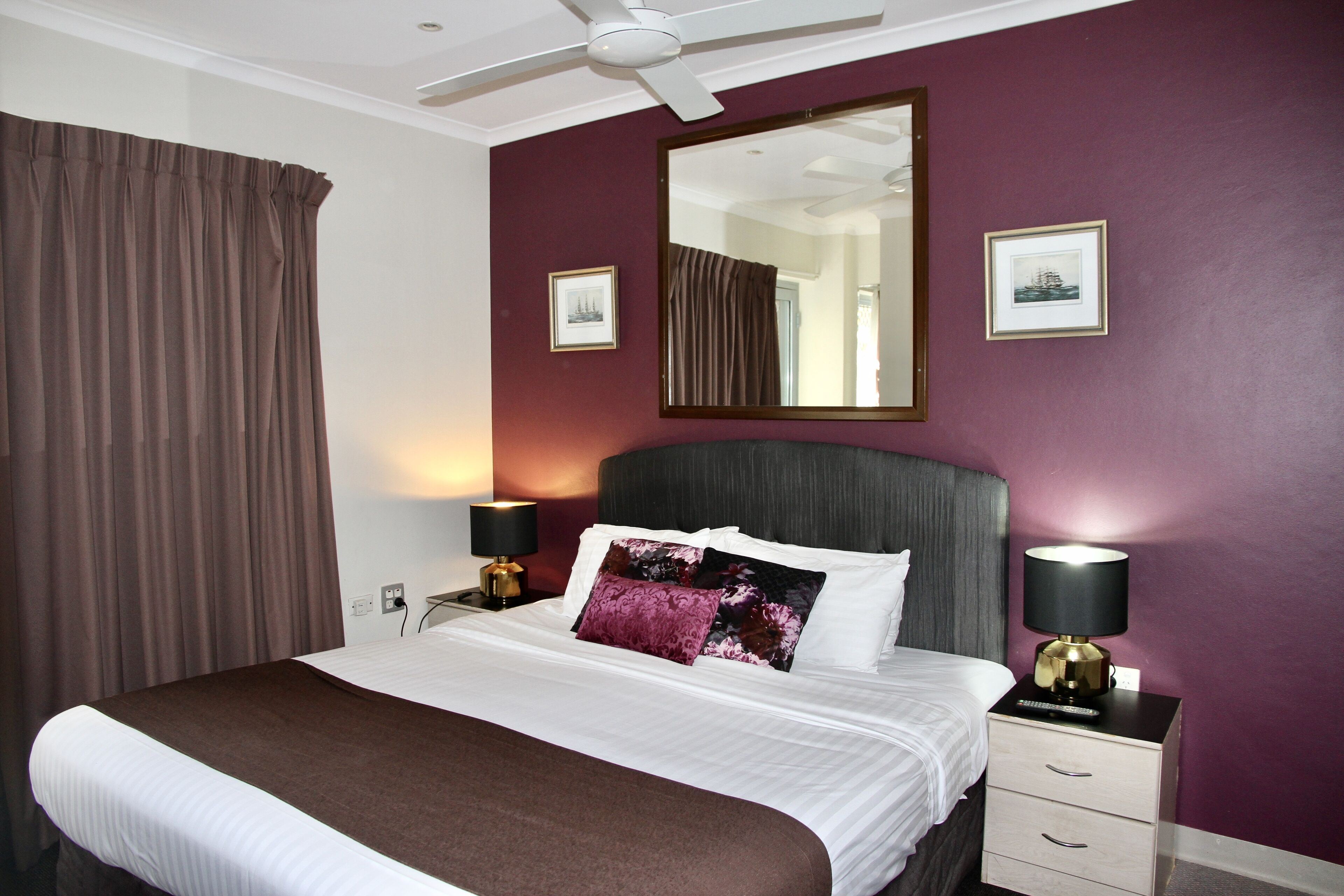 A hotel room with a bed, two bedside tables, a ceiling fan, a large mirror, and two framed pictures on the wall.