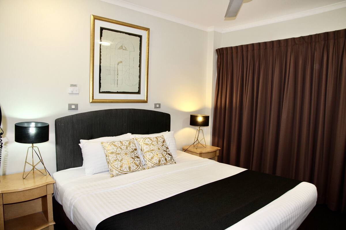 A hotel room with a large bed, two bedside lamps, a framed artwork on the wall, and a brown curtain.