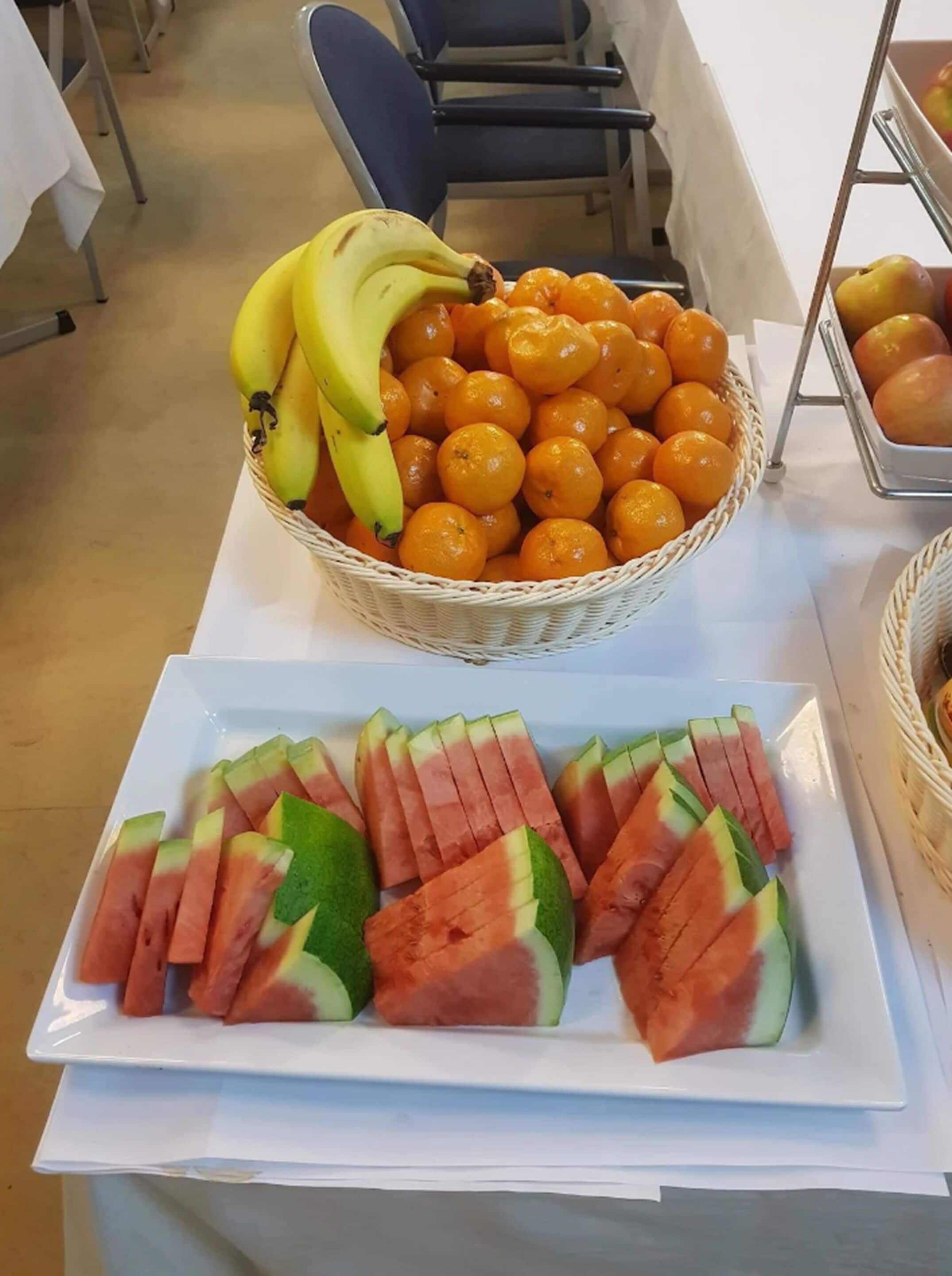 A table with a basket of oranges and bananas, and a plate of watermelon slices.