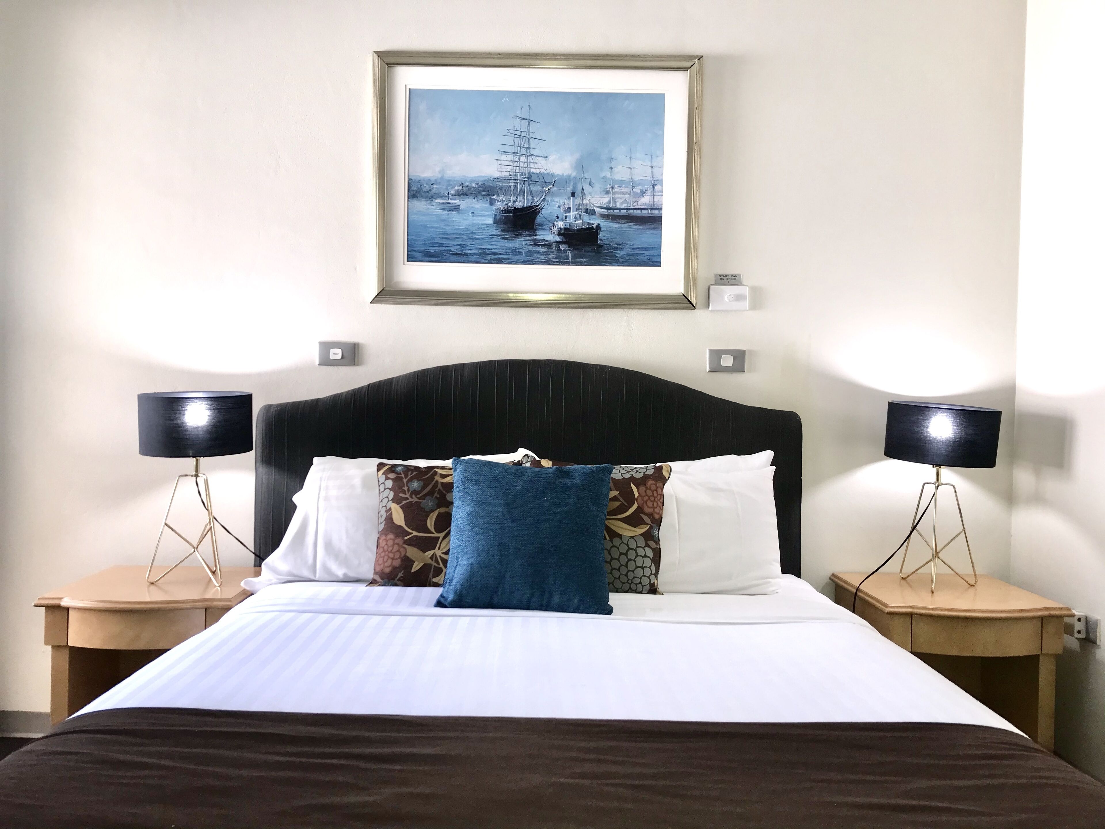 A neatly made bed with a white and brown bedspread, flanked by two nightstands with black lamps. Above the bed, there is a framed picture of a maritime scene.