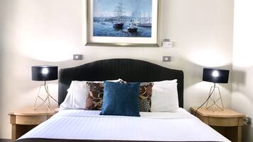 A neatly made bed with a white and brown bedspread, flanked by two nightstands with black lamps. Above the bed, there is a framed picture of a maritime scene.