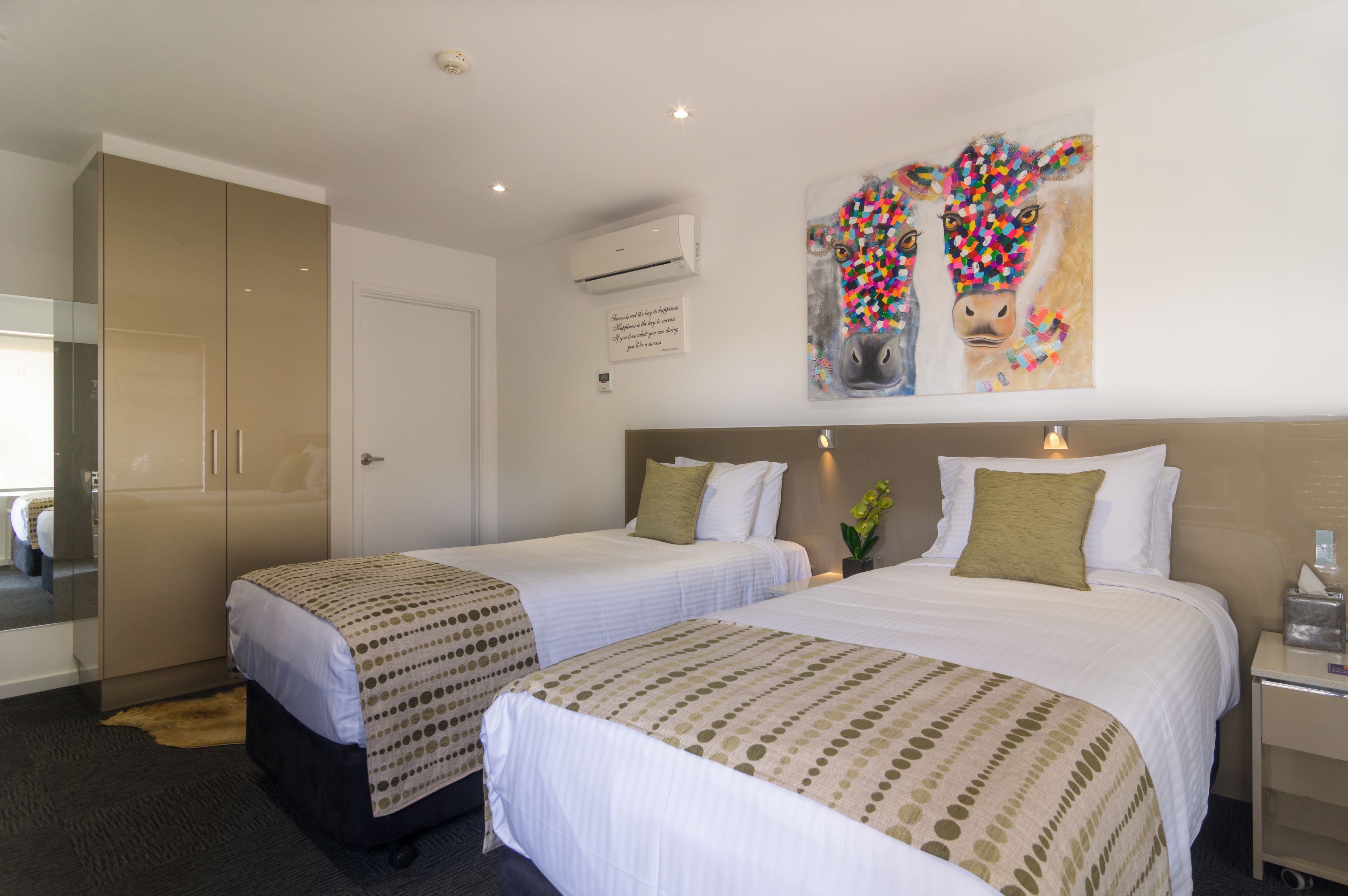 A hotel room with two beds, a large wardrobe, an air conditioning unit, and a colorful artwork on the wall.