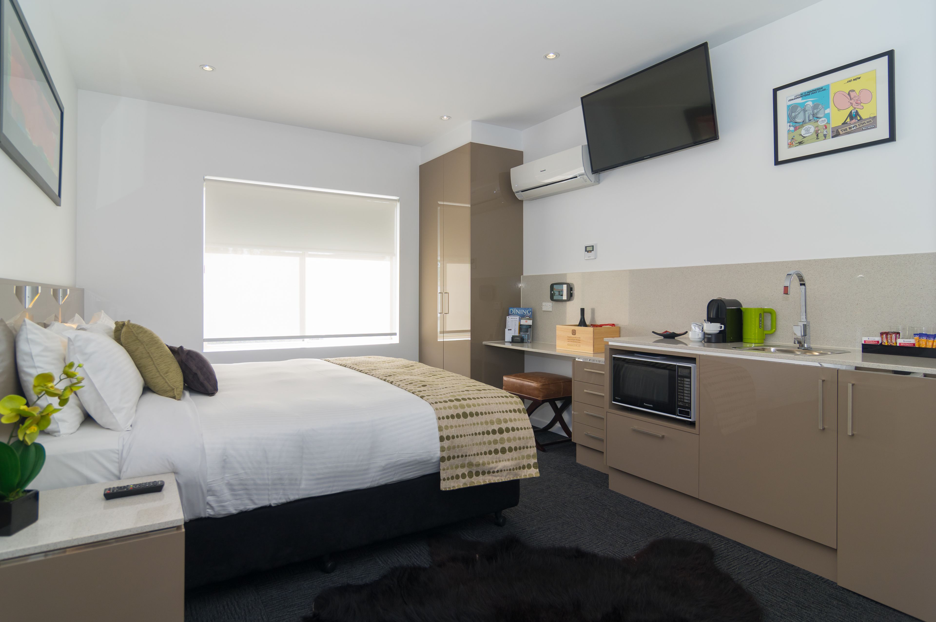 Ein modernes Hotelzimmer mit einem großen Bett, einem Schreibtisch mit Computer, einer Küchenzeile und einem an der Wand befestigten Fernseher.