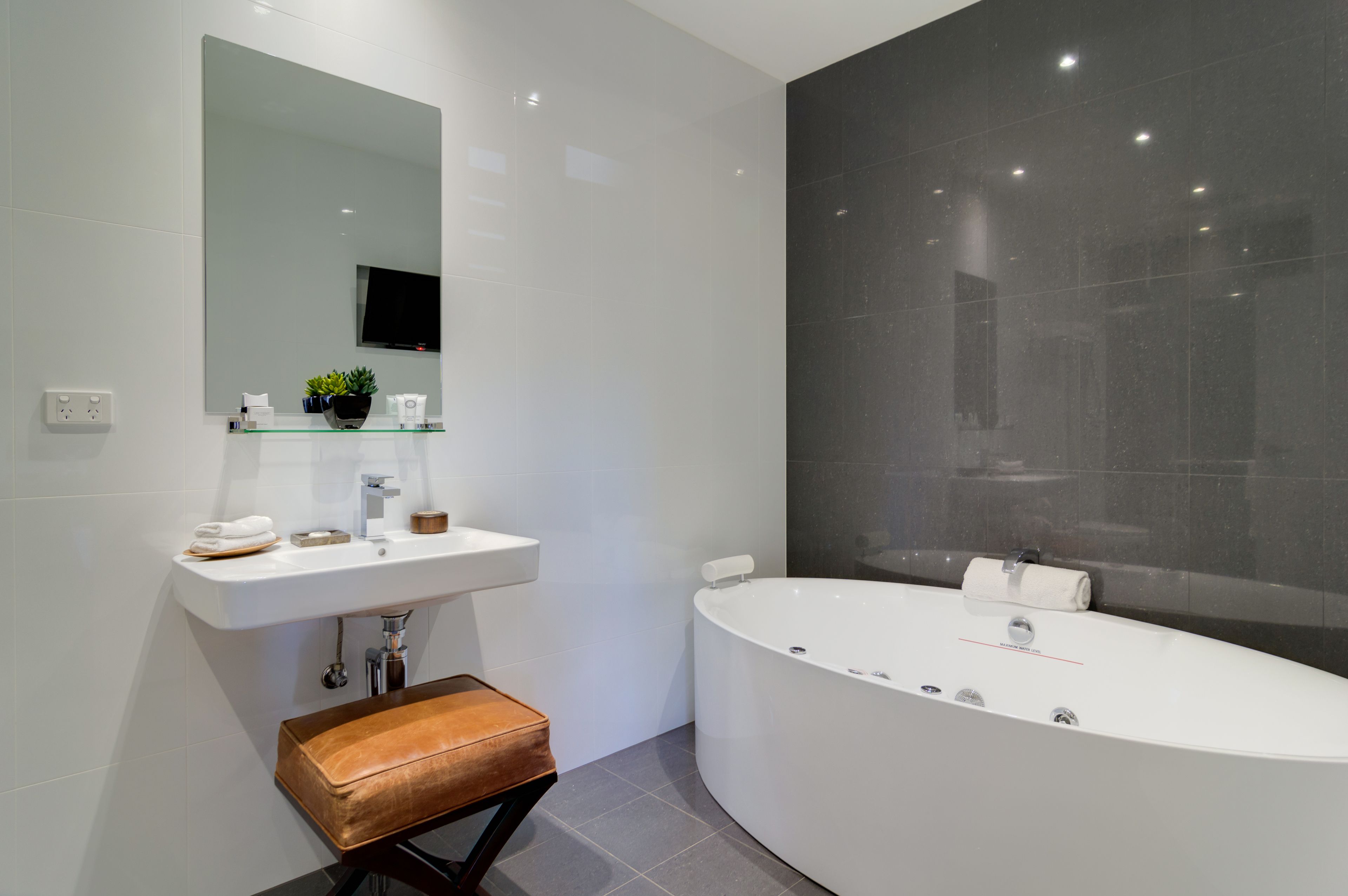 A modern bathroom with a white sink, a large mirror, a dark tiled wall, and a freestanding bench.