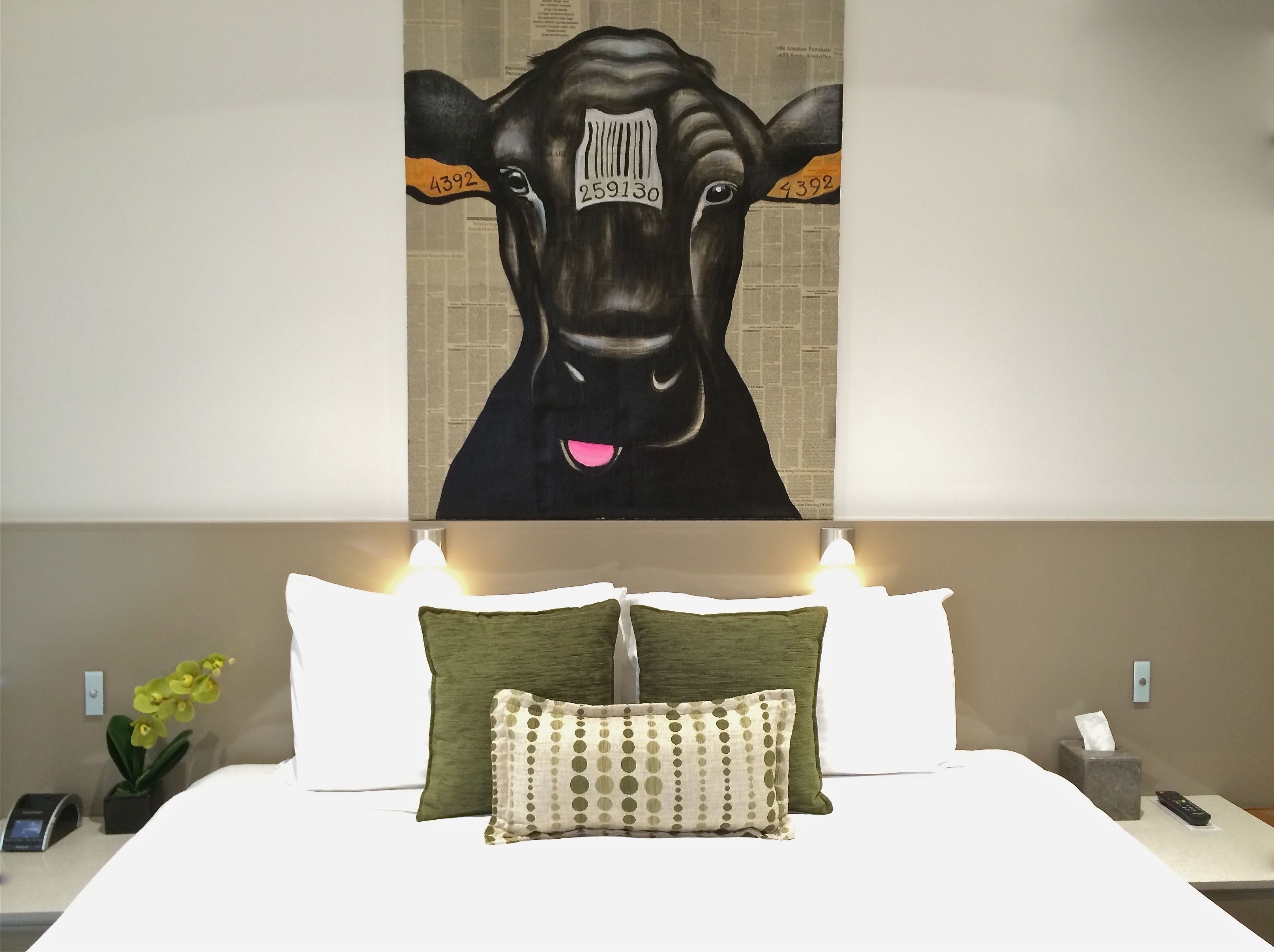 A hotel room with a large painting of a cow, a bed with white and green pillows, and bedside tables with a remote control and a tissue box.