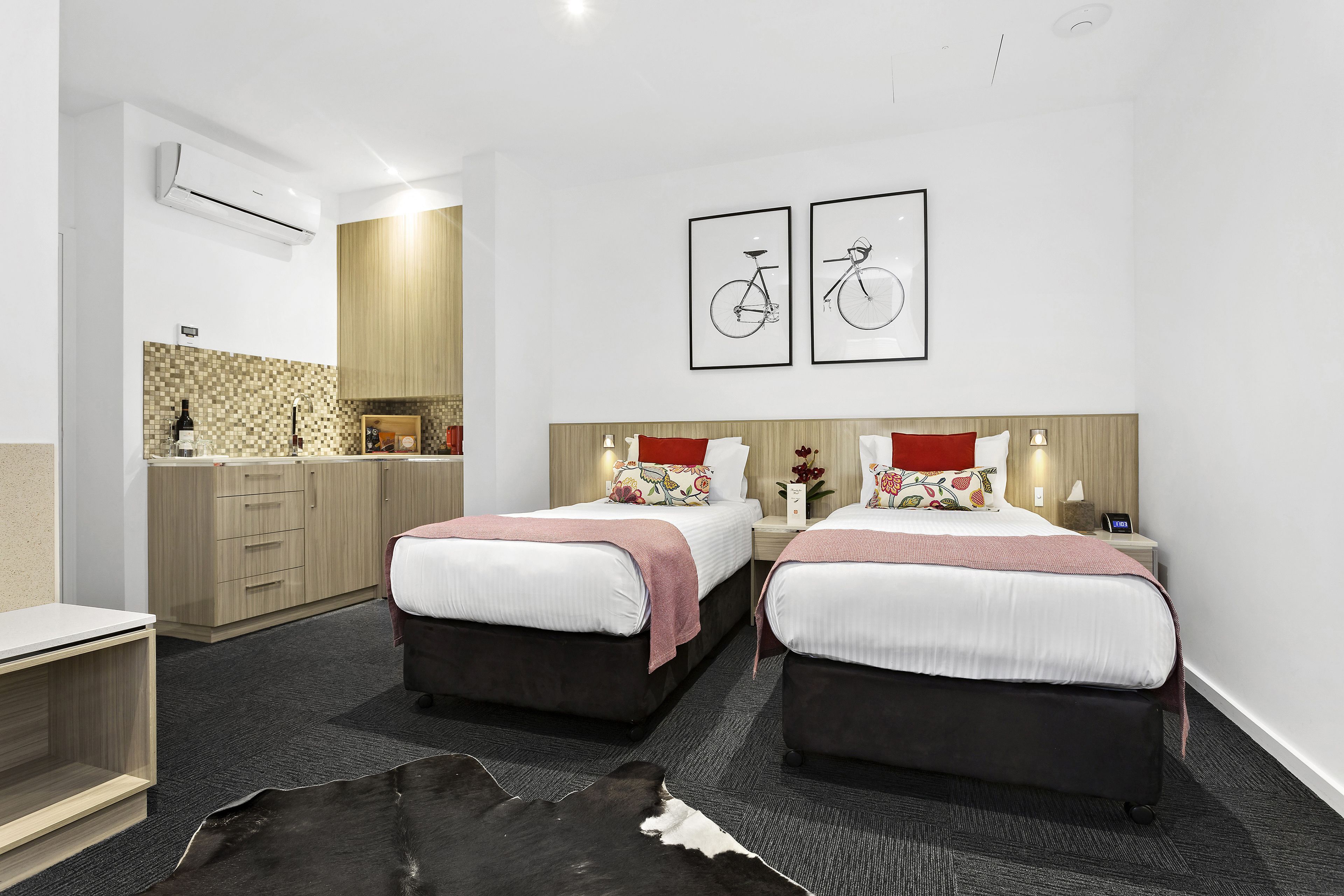 A hotel room with two beds, a small desk, a kitchenette, and a large mirror.