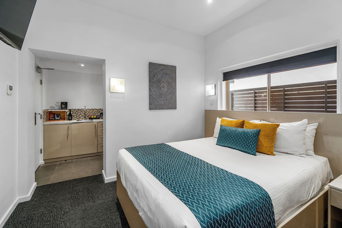 A hotel room with a bed, a window with blinds, a small kitchenette, and a bathroom.