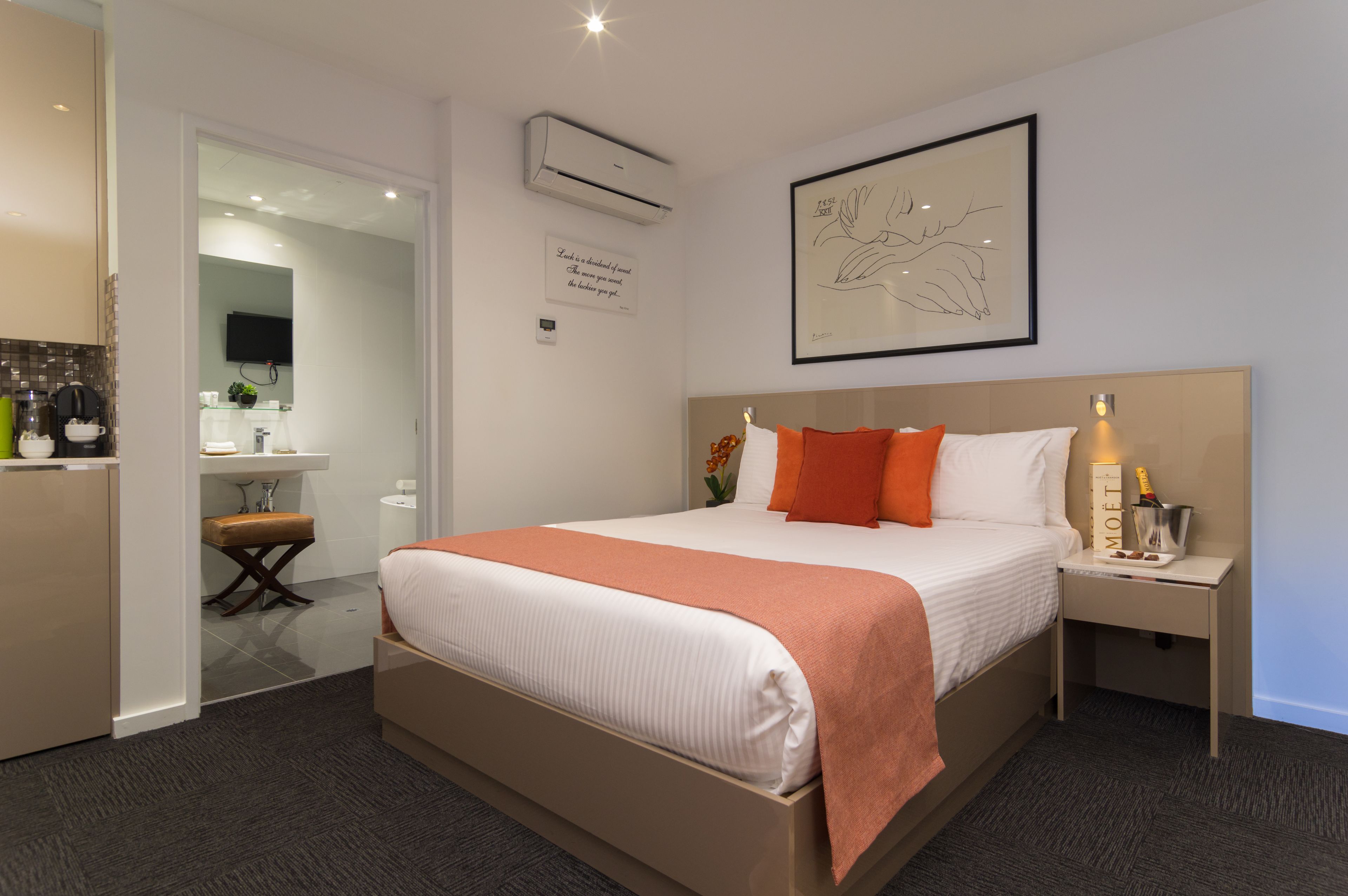 A hotel room with a bed, bedside table, nightstand, and a small desk with a chair. There is a wall-mounted air conditioner and a framed artwork above the bed.
