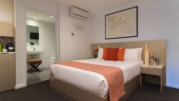 A hotel room with a bed, bedside table, nightstand, and a small desk with a chair. There is a wall-mounted air conditioner and a framed artwork above the bed.