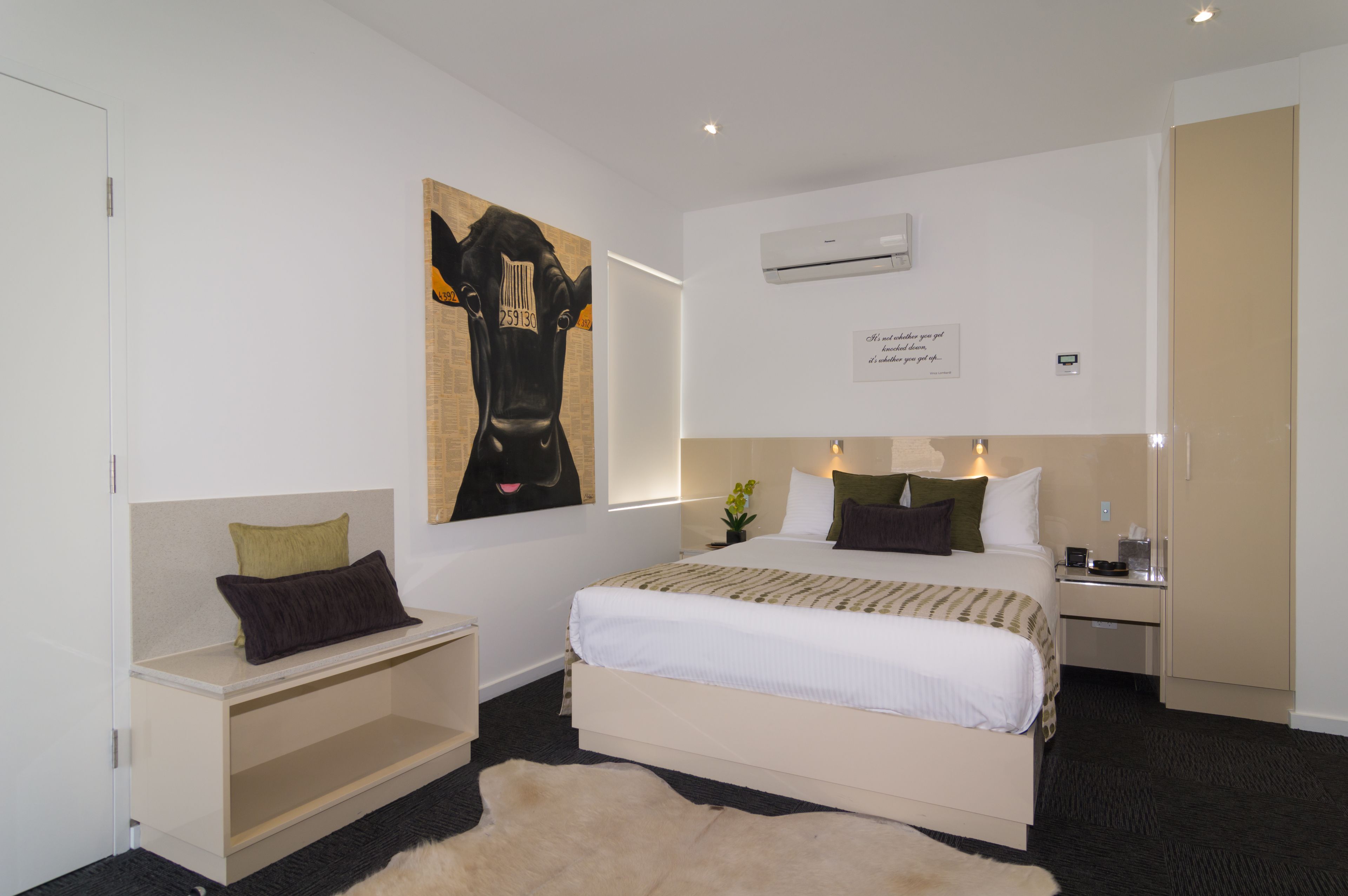 A hotel room with a large bed, bedside tables, a nightstand, a wall-mounted air conditioner, and a large artwork of a cow.