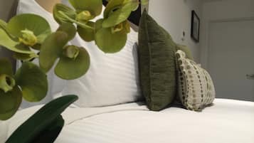 Uma cama bem arrumada com lençóis brancos, almofadas verdes e estampadas, e um vaso com uma orquídea verde em primeiro plano.
